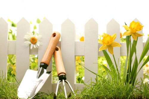 A close-up view of a small garden area in Millbank, featuring a white picket fence in the background, with bright yellow daffodils and white daisies emerging from the flower bed. In the foreground, there is a patch of lush, green grass with a slightly uneven surface, bordered by soil and small plants. Leaning against the fence are gardening tools, including a hand trowel with a wooden handle and a garden fork, indicating recent or ongoing garden maintenance. The scene is illuminated by natural daylight, suggesting a clear weather day, typical of spring or early summer, and reflects an outdoor space cared for by local gardening services. The garden layout displays a neat division between the flower bed and grassy area, with a simple paved or mulched section visible behind the plants, creating a balanced and tidy outdoor environment appropriate for residential landscaping in Millbank.