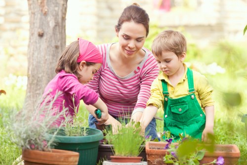 A woman engaging in gardening activities outdoors in a well-maintained garden in Millbank, surrounded by various potted plants, including herbs and flowering plants, with a lush green lawn, mature trees, and flower beds visible in the background. She is guiding two young children, one wearing a pink headscarf and the other in a green apron, as they work together on planting or tending to the garden beds, which are filled with vibrant, healthy foliage. The garden features a combination of grass, soil, and tiled or paved areas that provide a neat and accessible outdoor space for family activities and gardening. The natural lighting suggests a clear day, with sunlit areas enhancing the garden's colours. This scene reflects typical local outdoor gardening enhancements and the importance of family involvement in green spaces, consistent with gardening service offerings in the Millbank area.