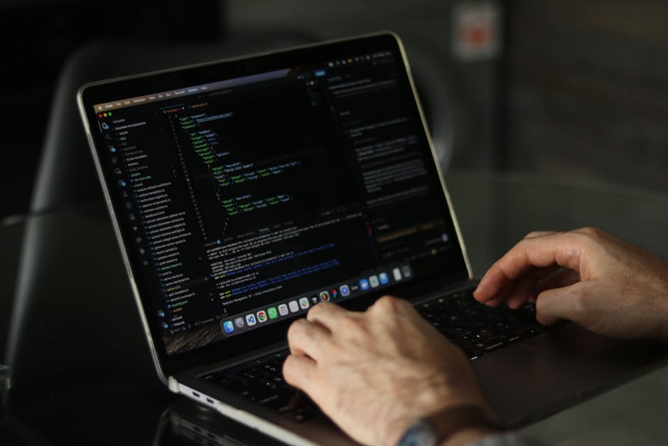 A pair of hands actively use a laptop placed on a dark surface, with the screen displaying lines of computer code in a dark-themed editor. The person's fingers rest on the keyboard, and the background