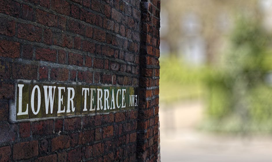 A close-up of a historic red brick wall featuring a rectangular metal street sign with white lettering reading 'LOWER TERRACE NW3,' mounted on the wall. The bricks have a rough texture with visible mo